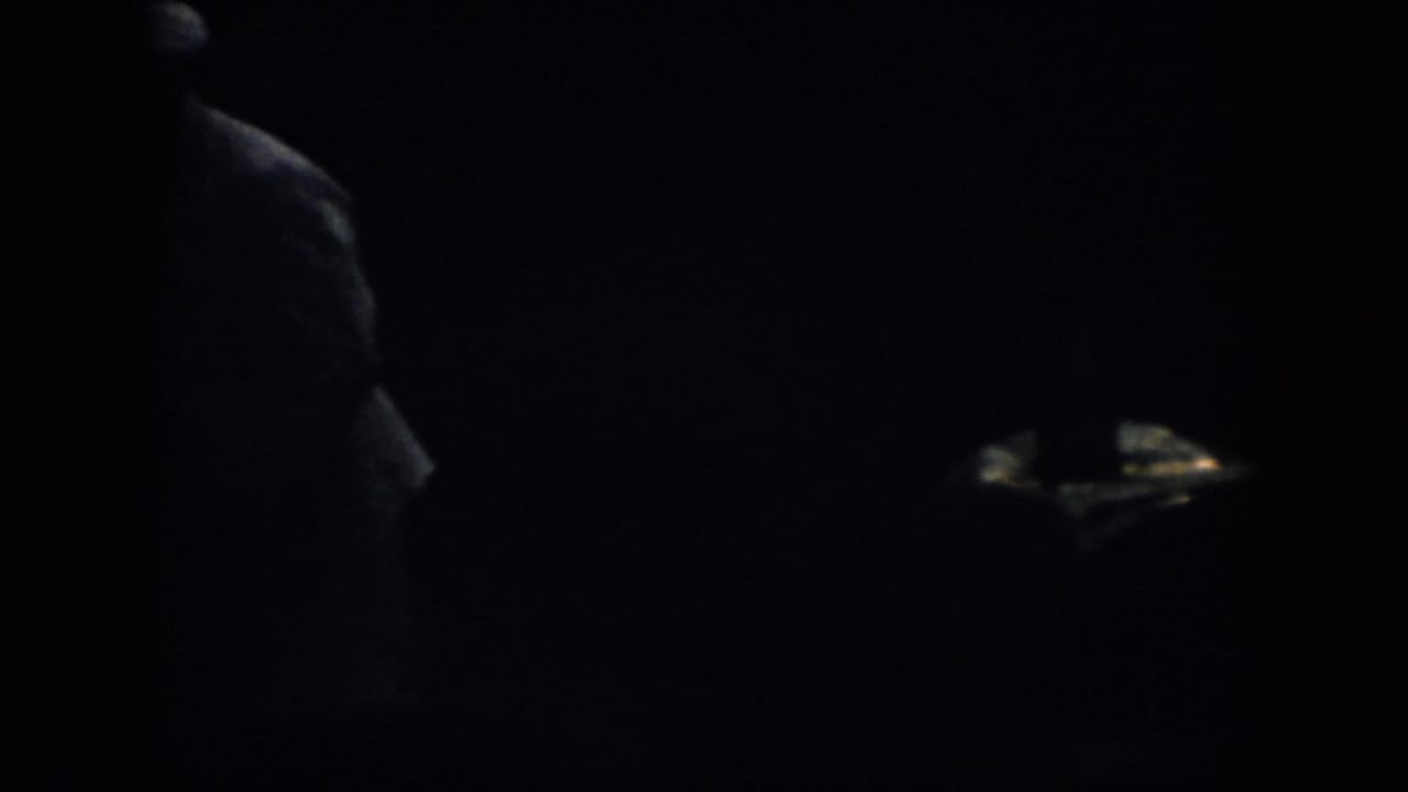 Very dark 1958 Virginia 8mm frame showing the side of a hat-wearing figure near the left edge, with most detail lost in shadow.
