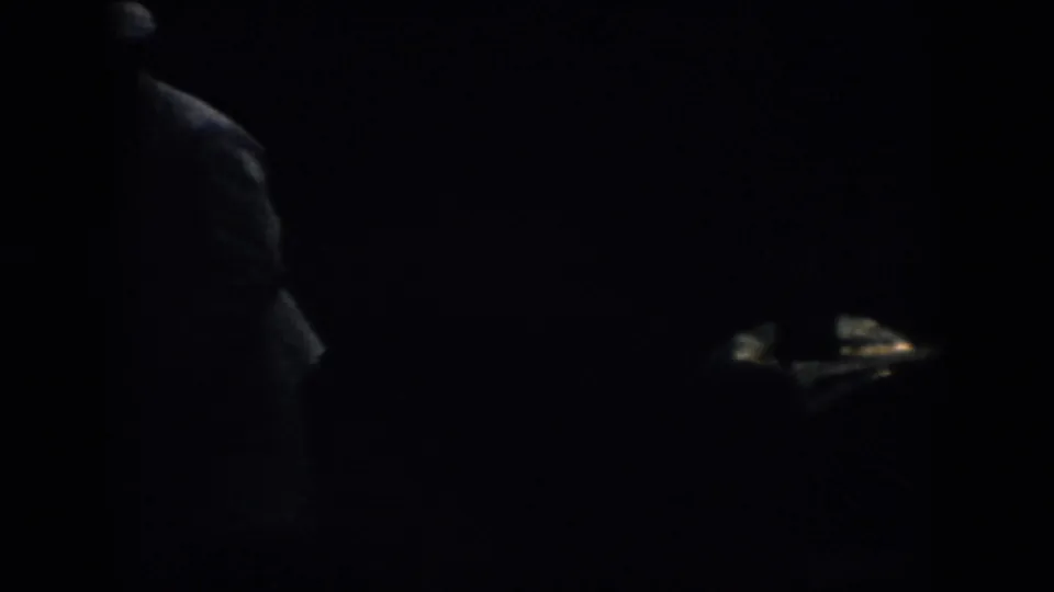 Very dark 1958 Virginia 8mm frame showing the side of a hat-wearing figure near the left edge, with most detail lost in shadow.
