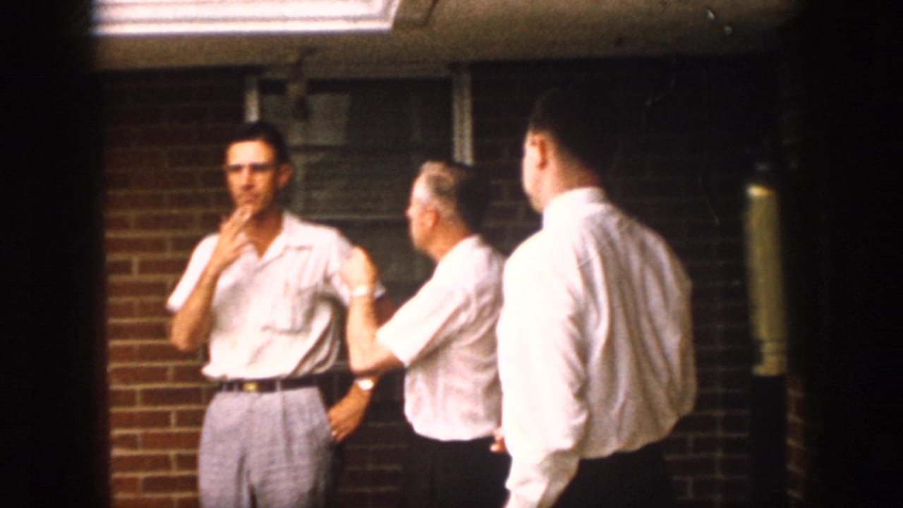 1958 Virginia 8mm frame showing three adult men in light shirts on a brick porch, one smoking and one wearing glasses near a window.