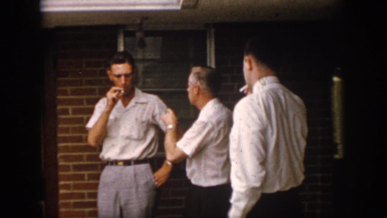 1958 Virginia home-movie frame of three adult men on a brick porch at night, including a smoking man facing the camera and another seen from behind.