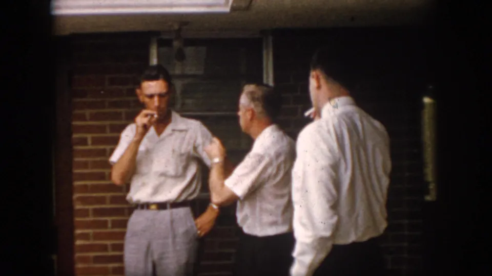 1958 Virginia home-movie frame of three adult men on a brick porch at night, including a smoking man facing the camera and another seen from behind.