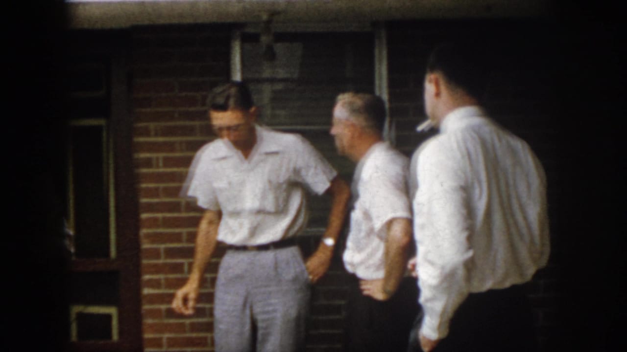 1958 Virginia home-movie frame of adult men in short-sleeved shirts talking beside a brick wall and dark paneled door.