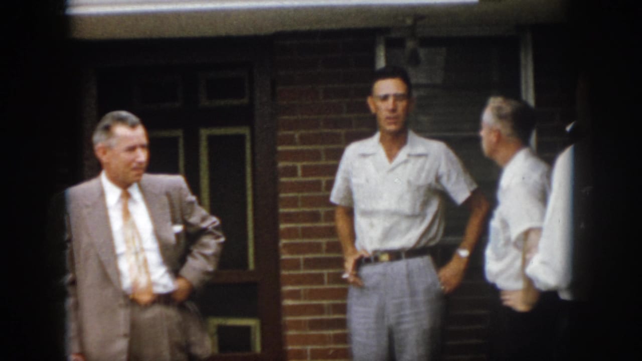 1958 Virginia porch scene with a suited man and two casually dressed men standing near a brick wall and dark doorway.