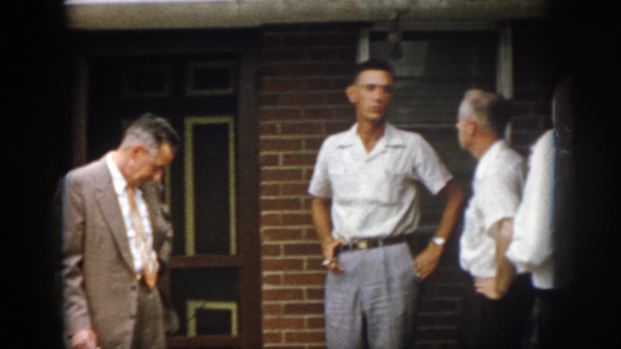 1958 Virginia home-movie still with a suited man looking down and casually dressed men gathered by a brick residential entrance.