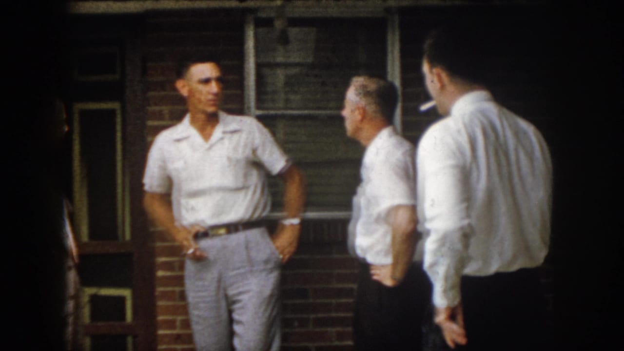 1958 Virginia frame of men conversing on a brick porch, with one man holding a cigarette near his waist.