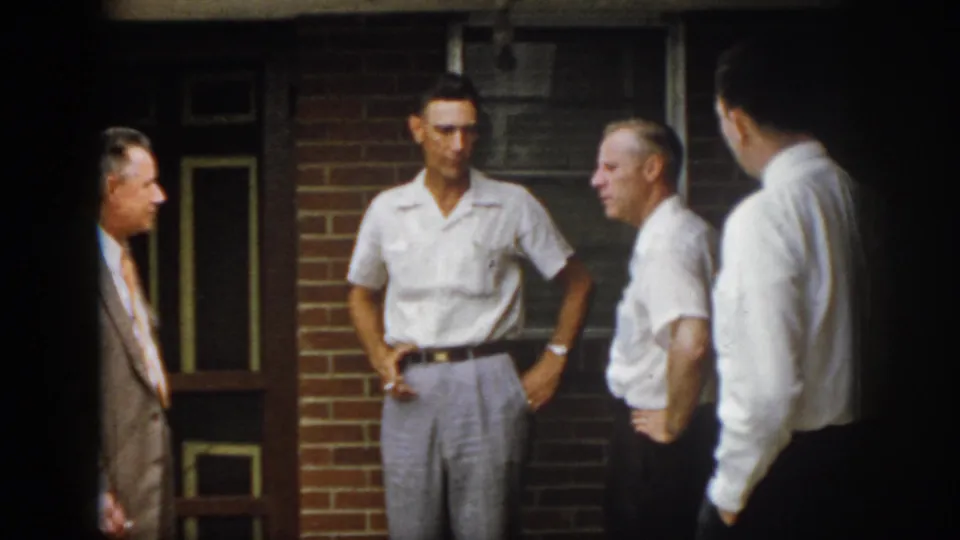 In 1958 Virginia home-movie footage, four adult men appear in conversation outside a brick building with windows and a doorway.