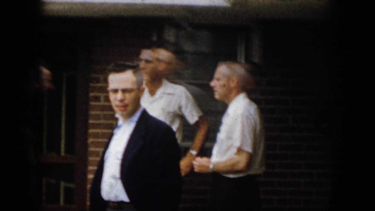 A man in a dark jacket faces the camera while other adult men stand near a brick building in 1958 Virginia footage.