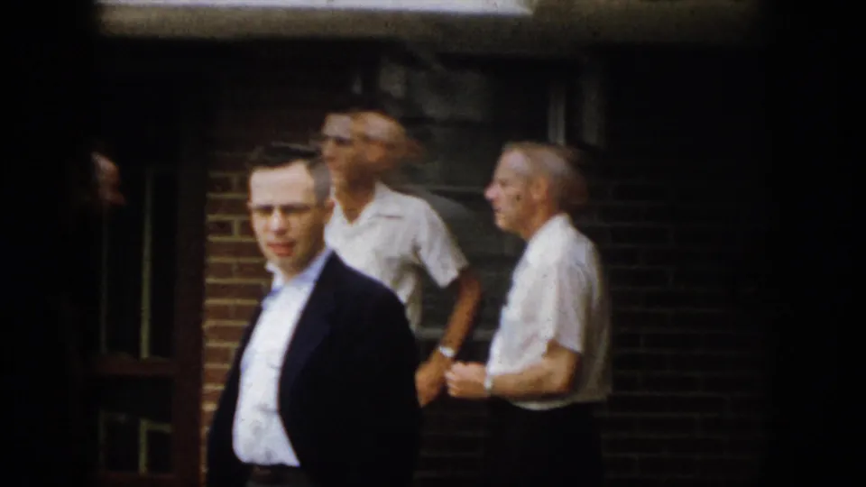 A man in a dark jacket faces the camera while other adult men stand near a brick building in 1958 Virginia footage.