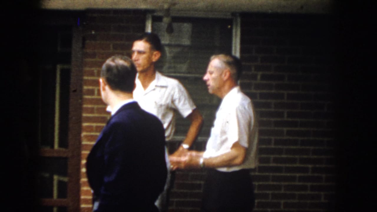 Adult men continue talking outside the brick building, with one man seen from behind in dark clothing and others in white shirts.