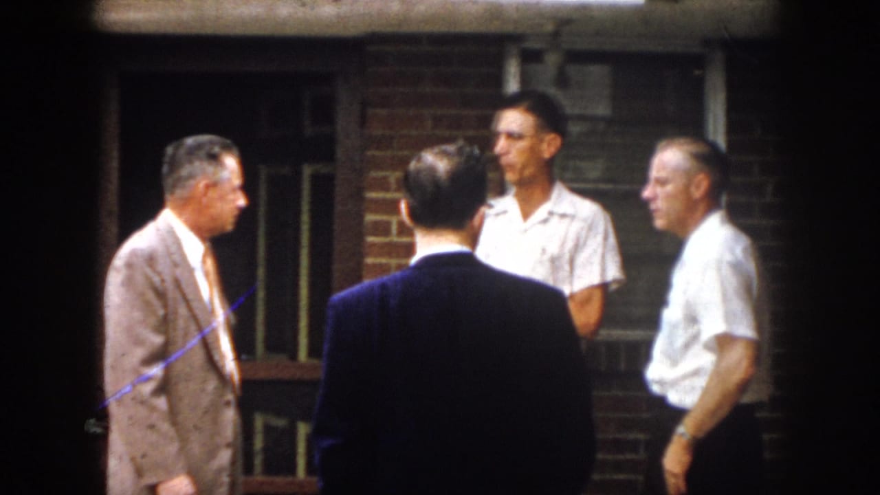 Four adult men stand in conversation outside a brick building, including one in a light suit and one viewed from behind in a dark jacket.