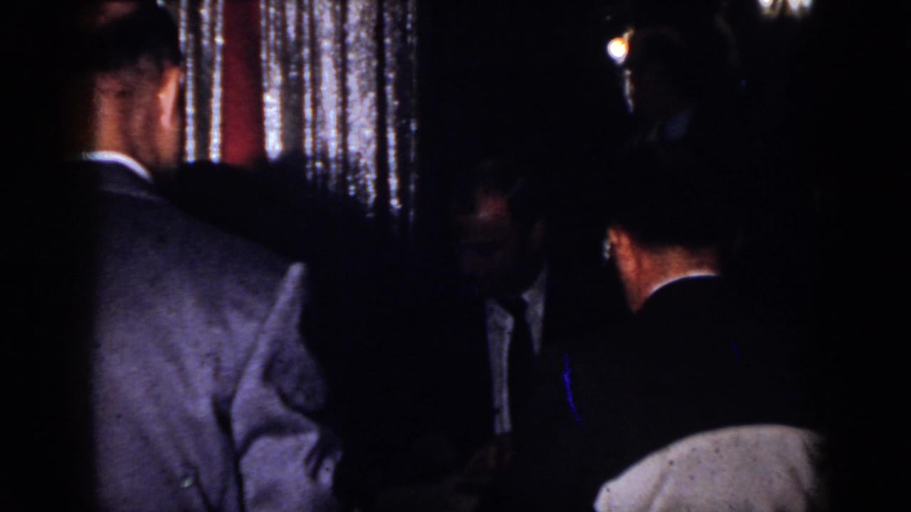 1958 Virginia home-movie frame showing adult men in suits conversing in a dark indoor party setting with shimmering curtains.