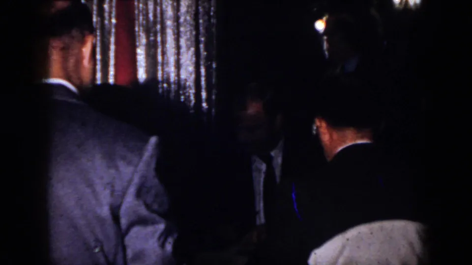 1958 Virginia home-movie frame showing adult men in suits conversing in a dark indoor party setting with shimmering curtains.