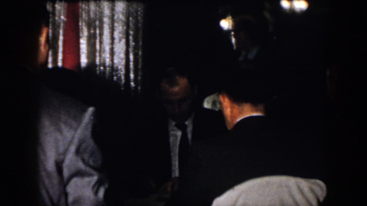 1958 Virginia indoor party frame with suited men gathered in low light, partially obscured by shadow and handheld framing.