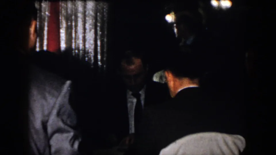 1958 Virginia indoor party frame with suited men gathered in low light, partially obscured by shadow and handheld framing.