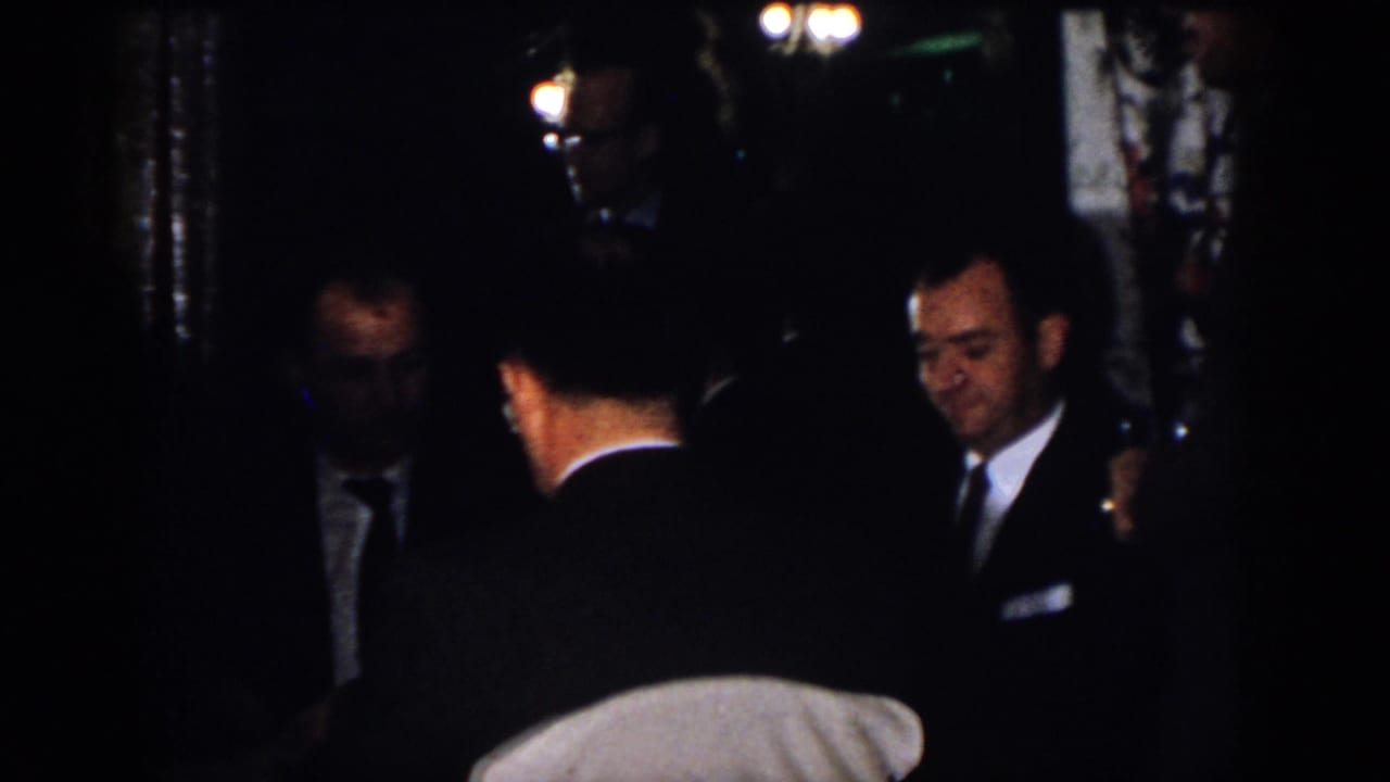 1958 Virginia home-movie still of adult men in suits talking indoors, including one face turned toward the camera.