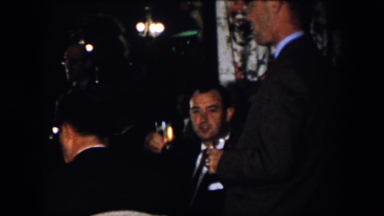 1958 Virginia indoor gathering frame showing suited men near chandeliers, including one man holding a drink.