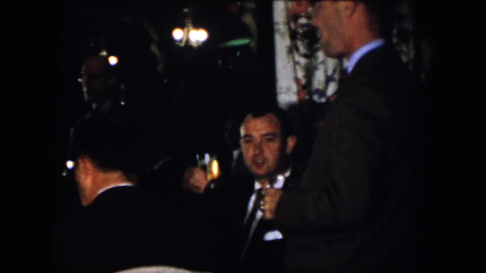 1958 Virginia indoor gathering frame showing suited men near chandeliers, including one man holding a drink.