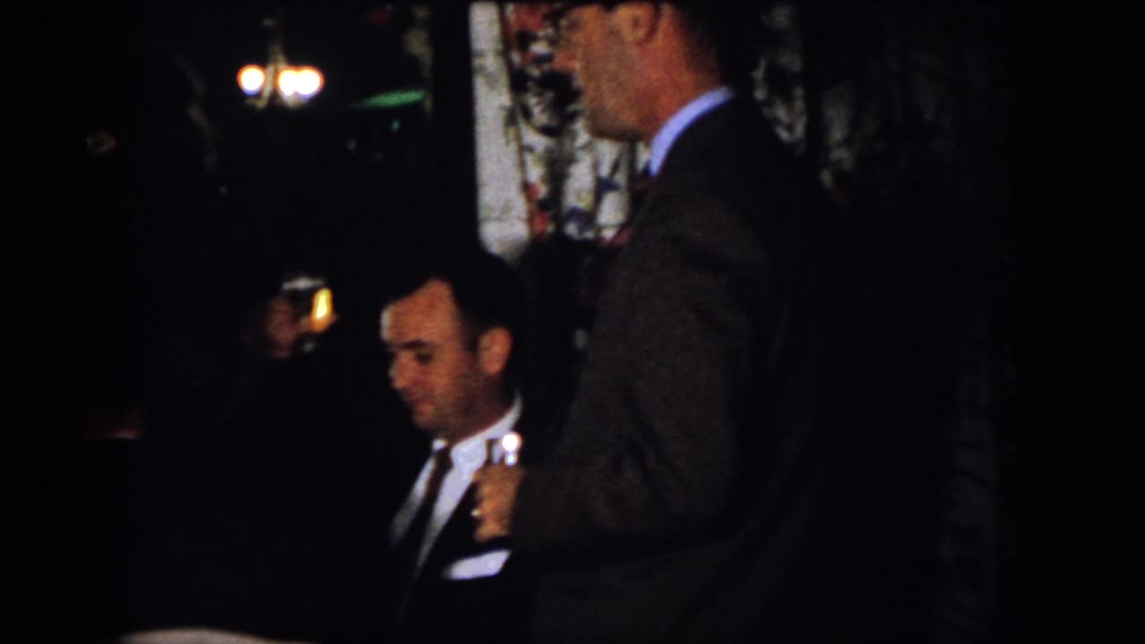 Men in suits sit and stand in a dim room, with one holding a drink.