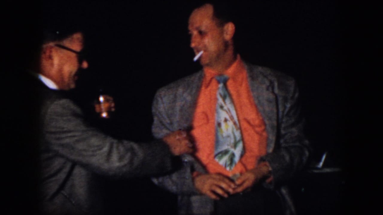A man with glasses reaches toward a smiling man in an orange shirt holding a cigarette.