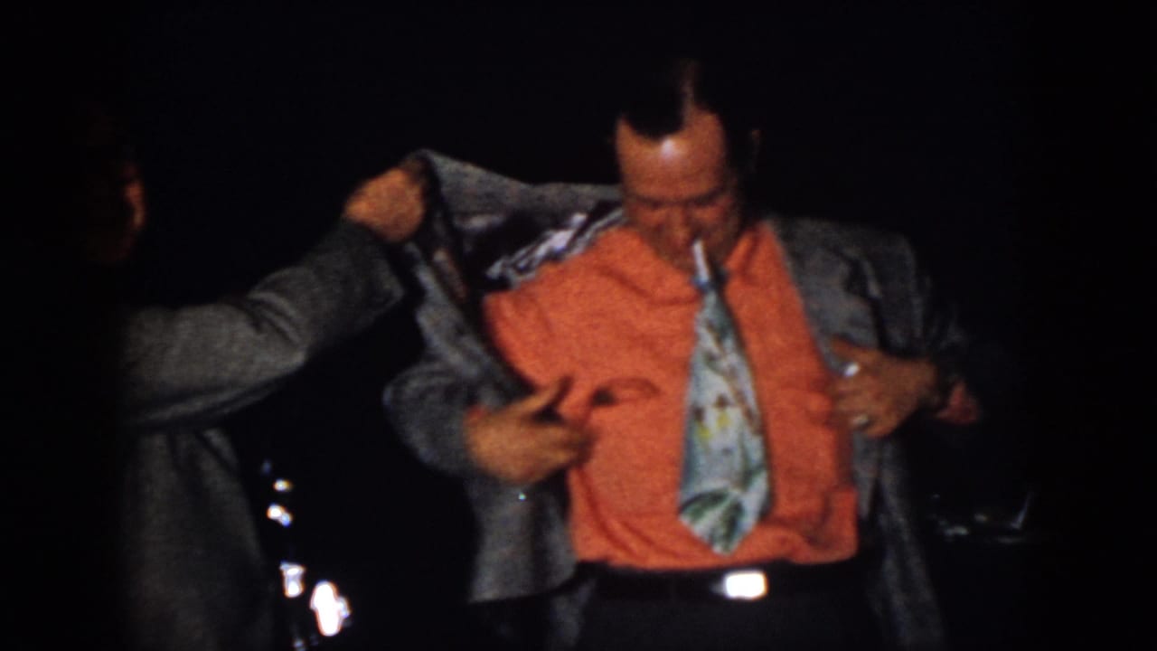 In the dim 1958 Virginia party scene, a man in an orange shirt and patterned tie looks down while his jacket is adjusted.