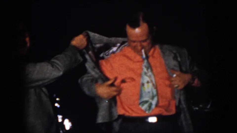 In the dim 1958 Virginia party scene, a man in an orange shirt and patterned tie looks down while his jacket is adjusted.