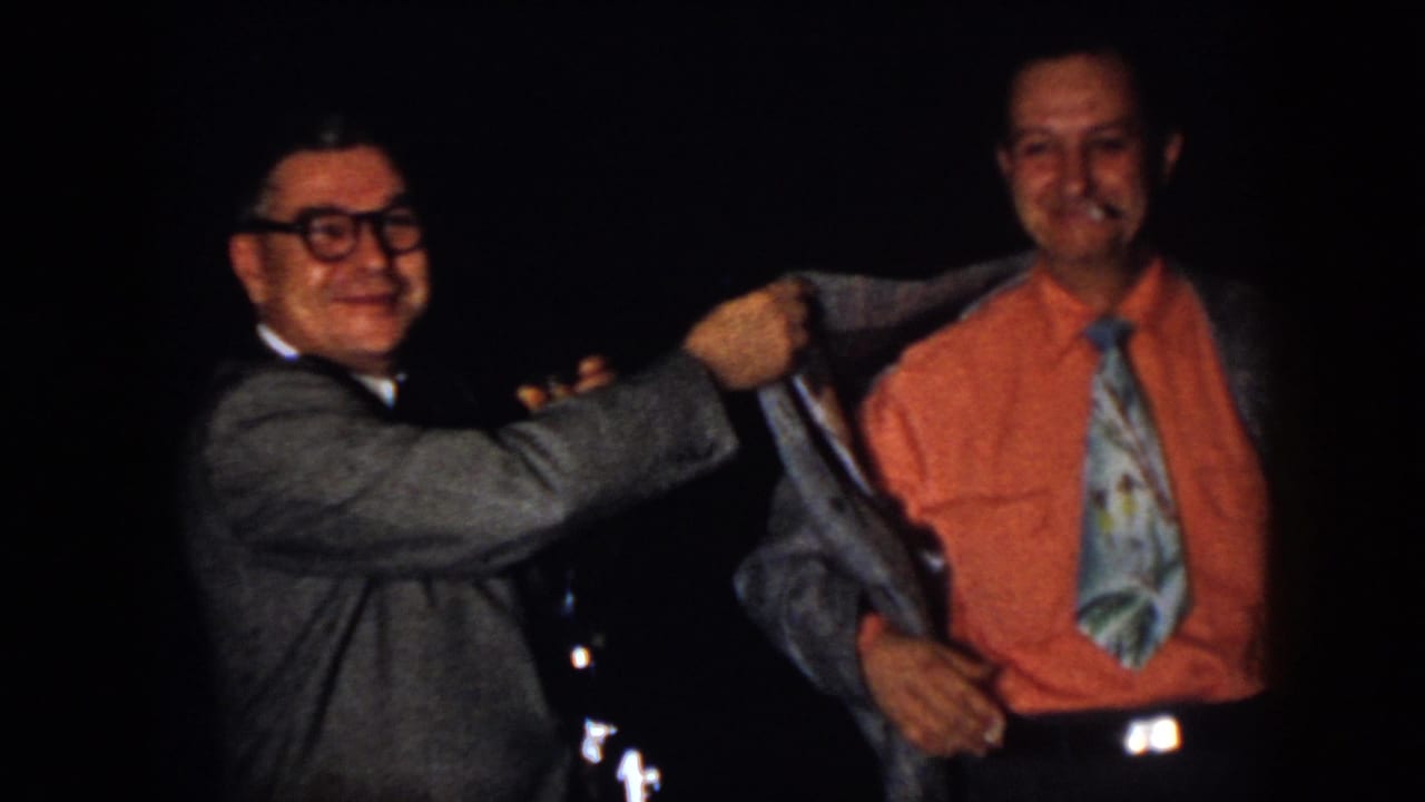 1958 Virginia home-movie frame with two men smiling during a playful jacket adjustment at an indoor party.