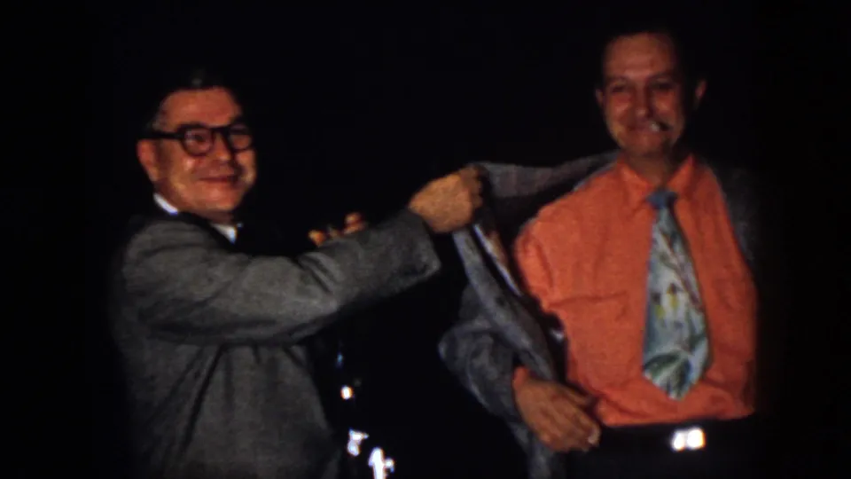 1958 Virginia home-movie frame with two men smiling during a playful jacket adjustment at an indoor party.
