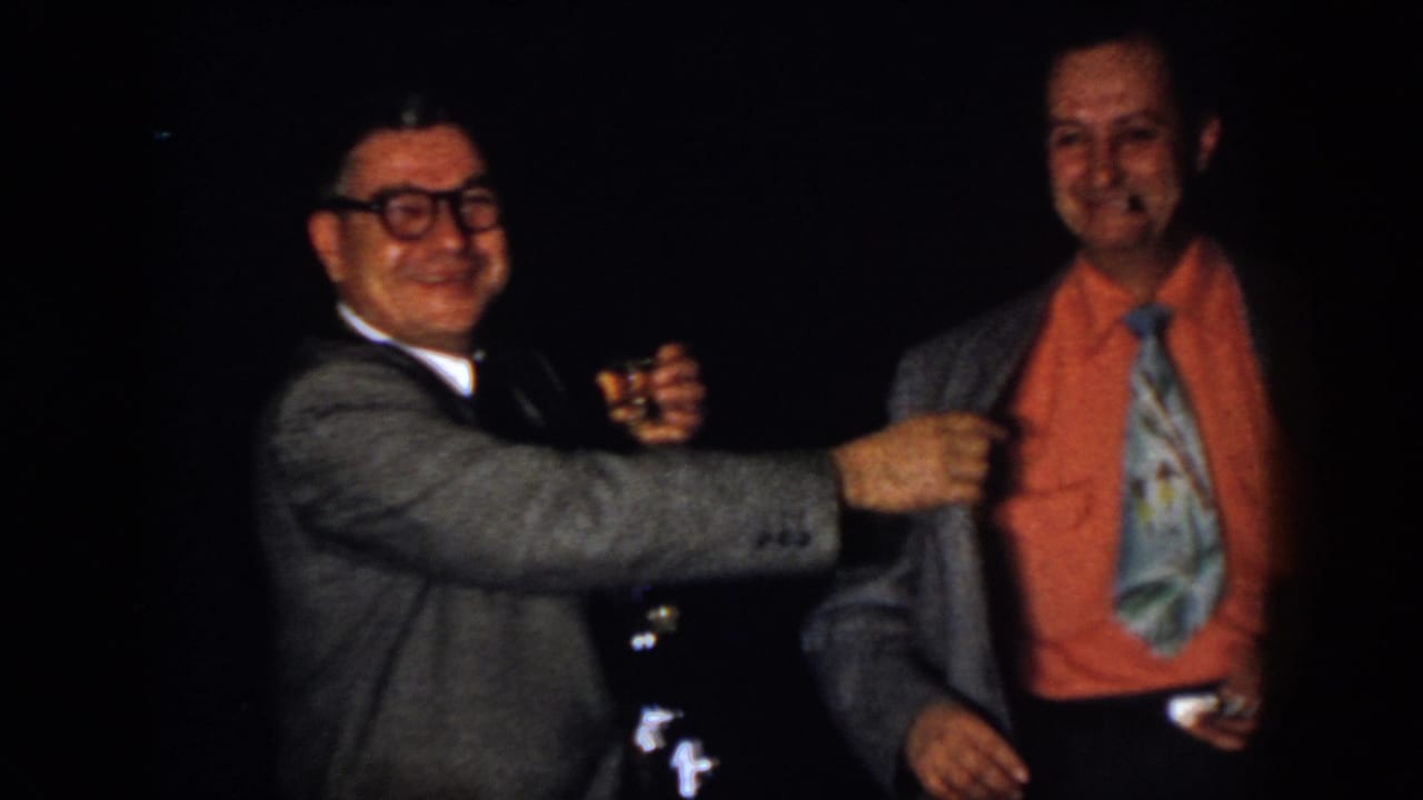 Two men in suits smile in a dark room, one wearing glasses and the other an orange shirt and patterned tie.