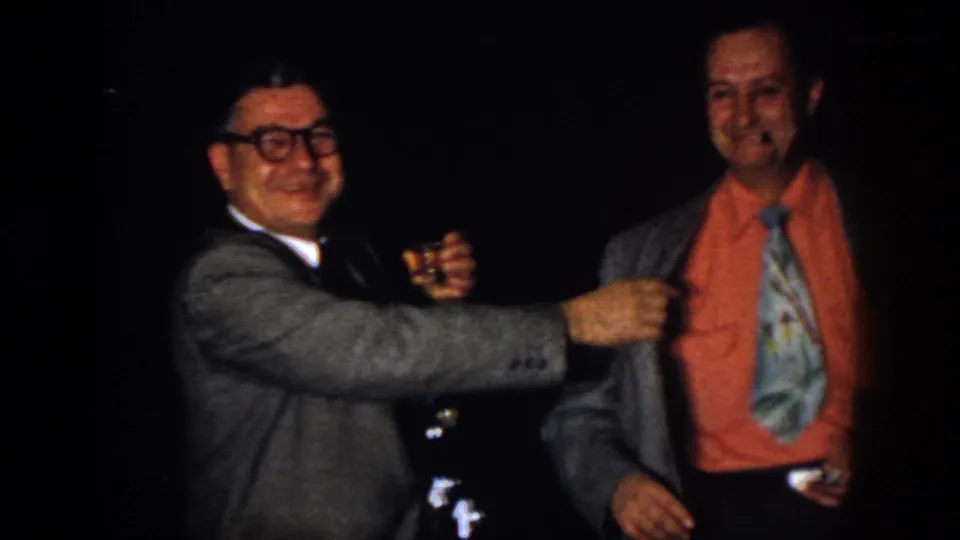 1958 Virginia home-movie frame of two adult men smiling indoors, one in glasses and a gray suit, the other in an orange shirt and patterned tie.