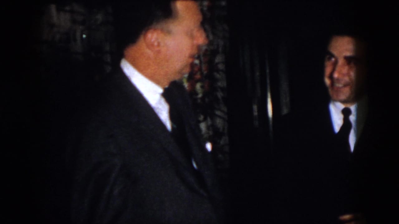 A man in a dark suit turns toward another smiling man in a dark interior.