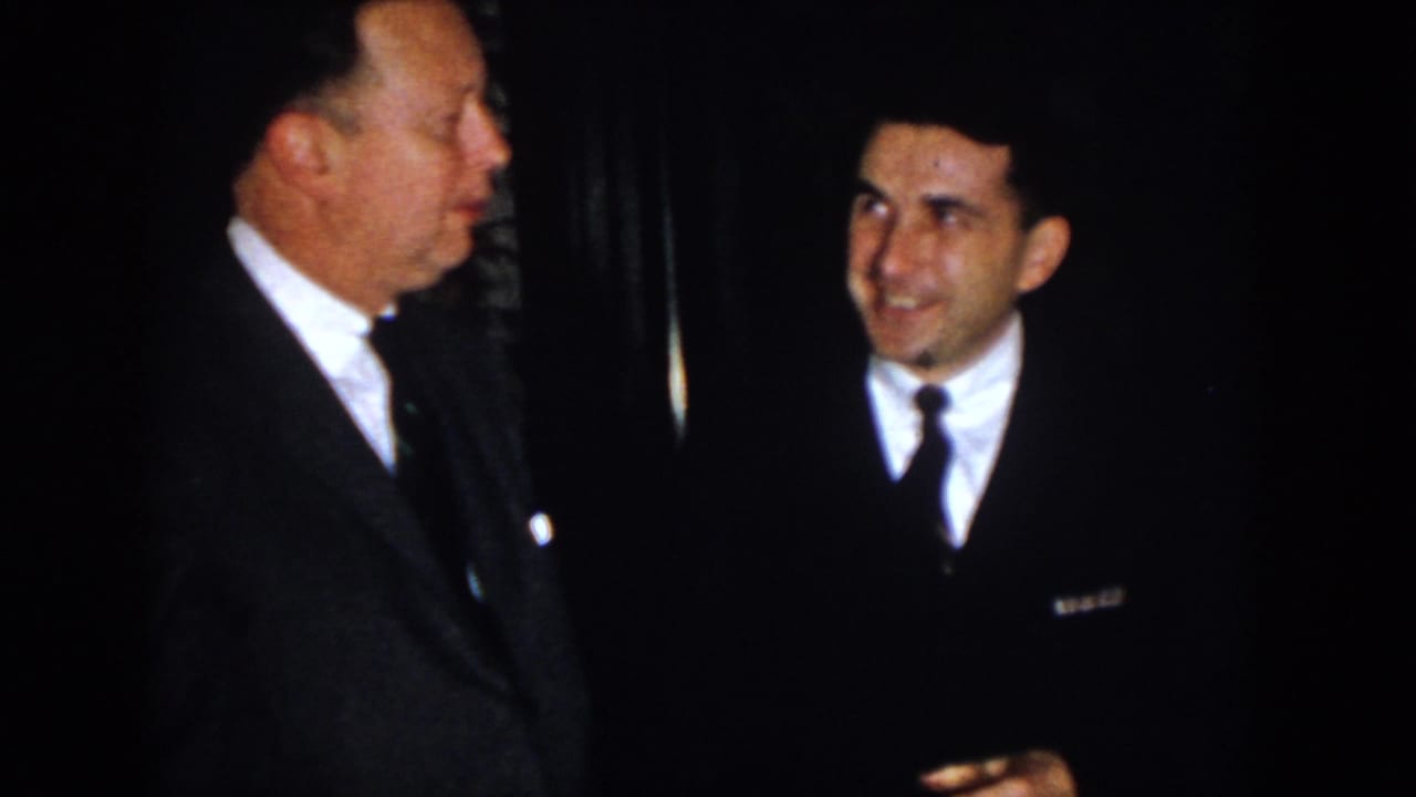 1958 Virginia home-movie frame of two suited adult men conversing indoors, one seen in profile and the other smiling in low light.