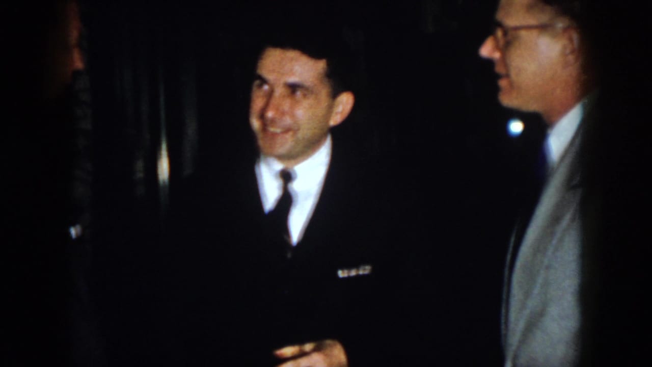 1958 Virginia frame of a smiling suited man in conversation with two nearby adult men at an indoor gathering.