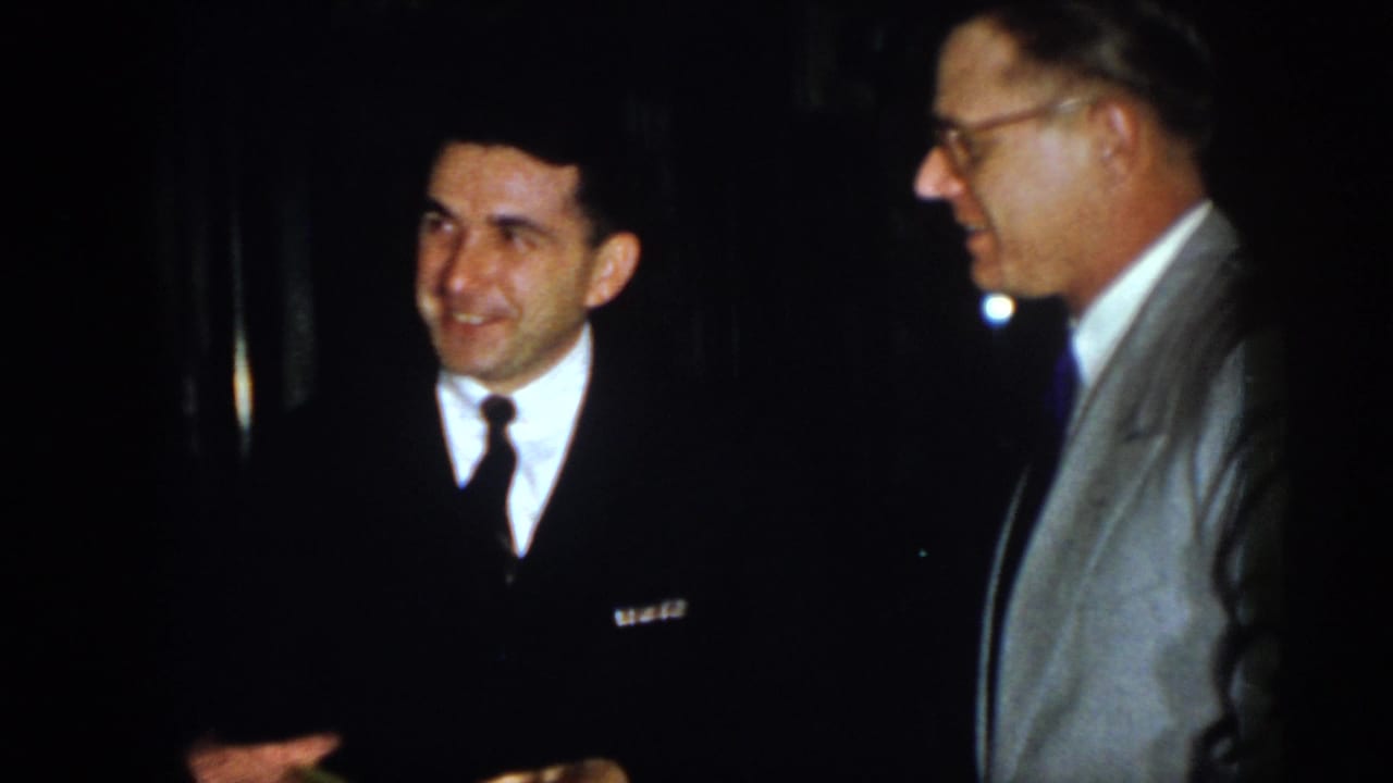 In this 1958 Virginia home-movie frame, a man in a dark suit and narrow tie smiles while a bespectacled man in a light gray jacket stands nearby in a dark indoor setting.