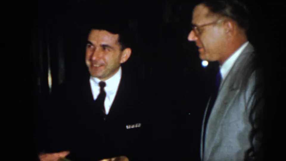 In this 1958 Virginia home-movie frame, a man in a dark suit and narrow tie smiles while a bespectacled man in a light gray jacket stands nearby in a dark indoor setting.