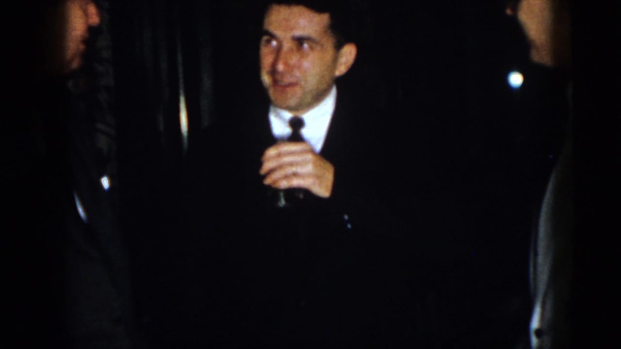 Man in a dark suit holds a small drink glass in a dim room.