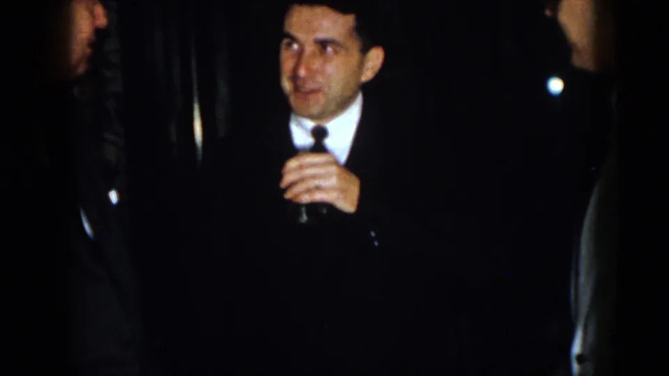 A man in a dark suit and narrow tie, possibly wearing uniform-style insignia, holds a small drink glass during the 1958 Virginia social-event footage.