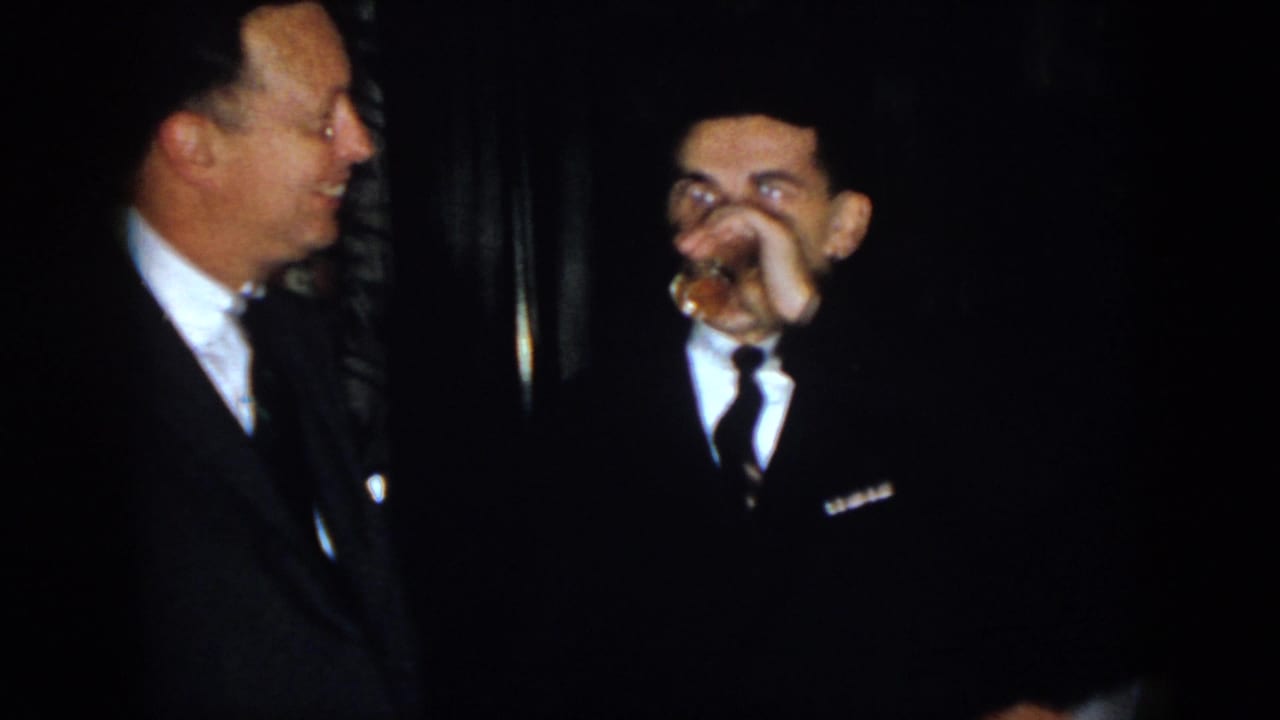 Two adult men in formal clothing laugh in a dim indoor space, with the man in the dark suit raising a small glass to his mouth.