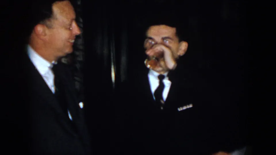 Two adult men in formal clothing laugh in a dim indoor space, with the man in the dark suit raising a small glass to his mouth.