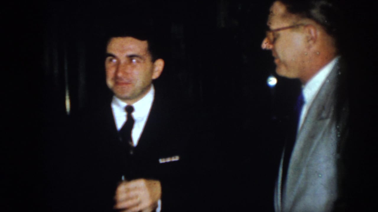 A man in a dark suit and narrow tie looks toward a bespectacled man in a light gray jacket, both standing close together at the dim 1958 Virginia gathering.