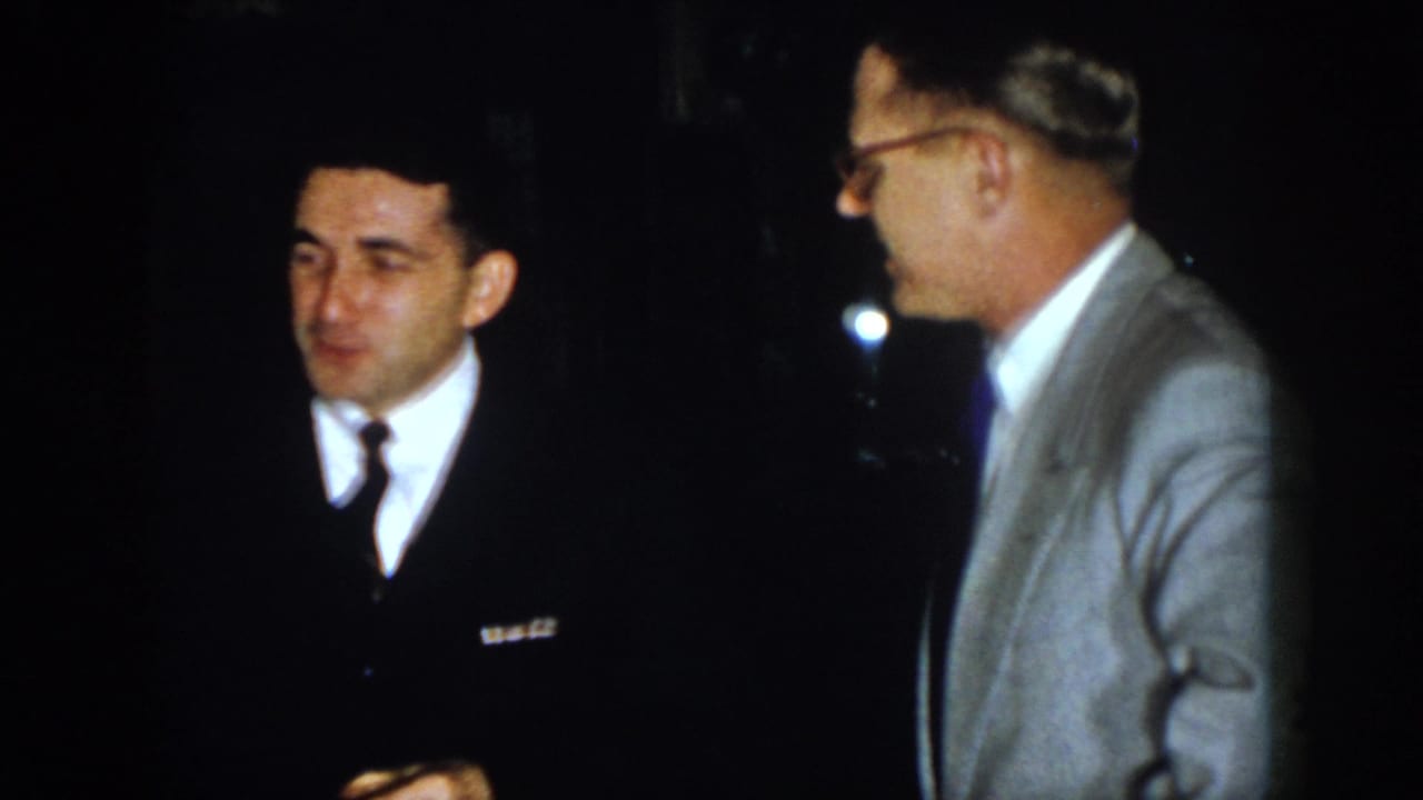 1958 Virginia home-movie frame showing suited adult men in low light, including one man in glasses and a light gray jacket.