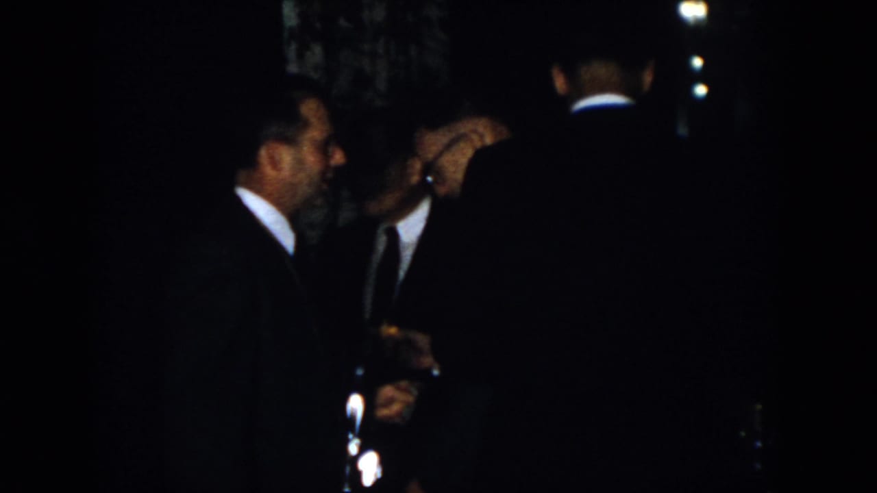 Dim 1958 Virginia home-movie frame of adult men in dark suits clustered together, partly obscured by shadows.