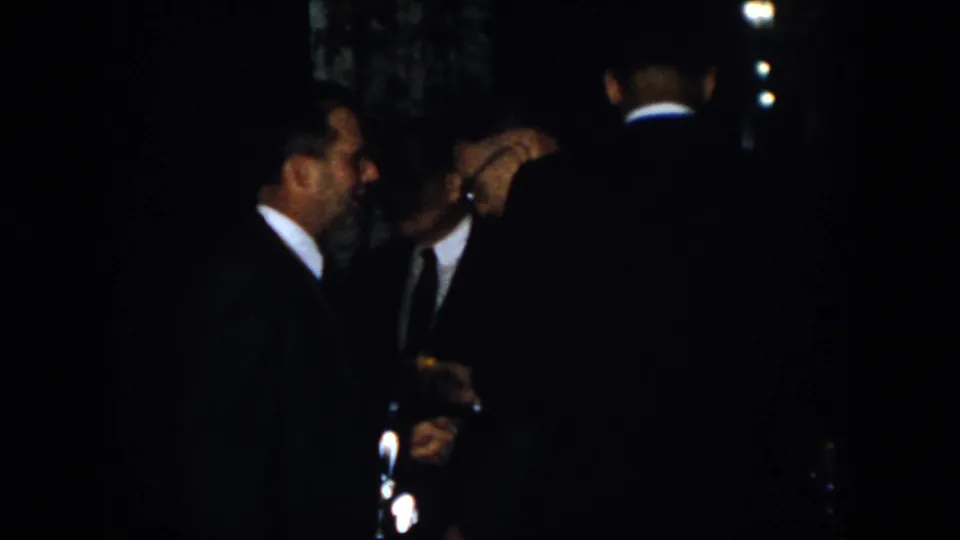 Dim 1958 Virginia home-movie frame of adult men in dark suits clustered together, partly obscured by shadows.
