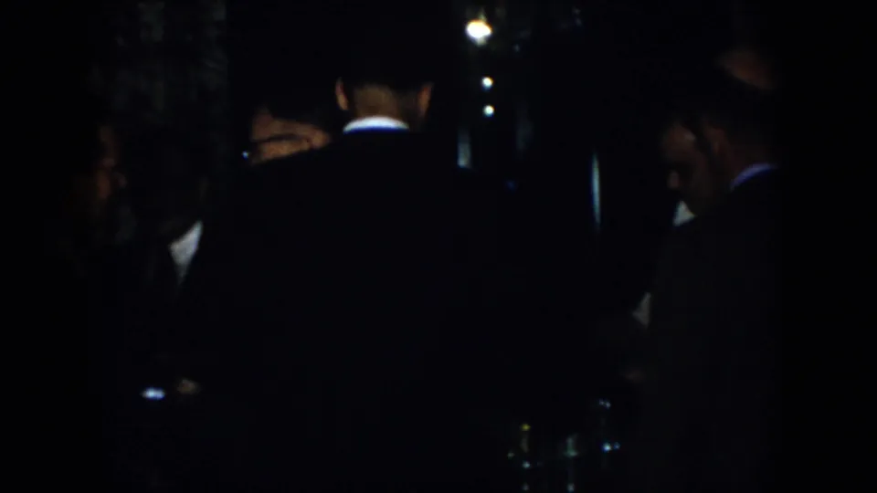 Low-light 1958 Virginia home-movie frame with suited adult men seen in partial silhouette during a crowded social moment.