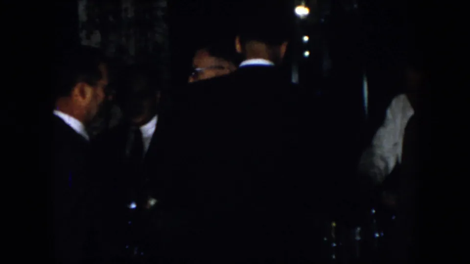 1958 Virginia 8mm frame showing adult men in formal suits conversing in a dim setting, with faces partly hidden by shadow.