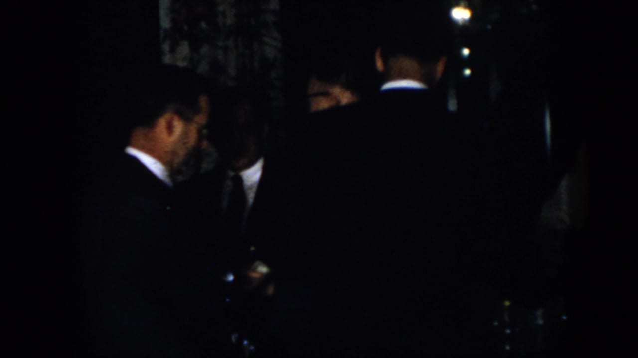 1958 Virginia home-movie frame showing several suited men conversing in a dark indoor room.