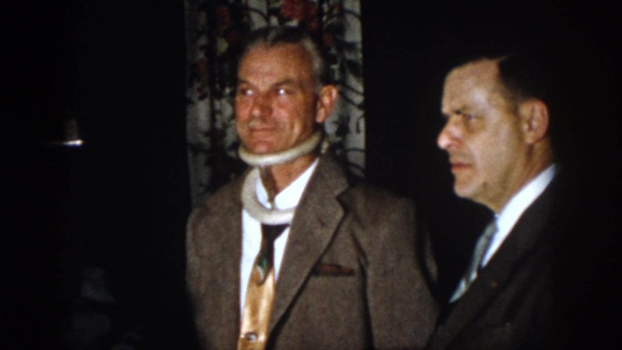 1958 Virginia frame of a suited man in a white neck brace standing near another man in a formal interior.