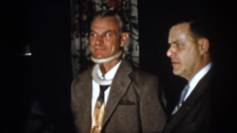 1958 Virginia frame of a suited man in a white neck brace standing near another man in a formal interior.