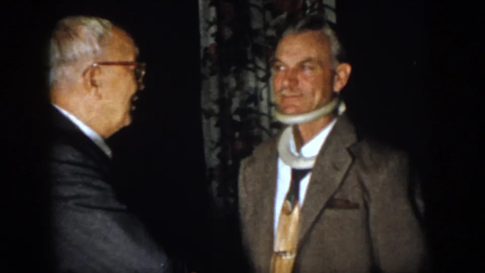 1958 Virginia home-movie frame showing an older man in glasses speaking with a suited man wearing a neck brace.