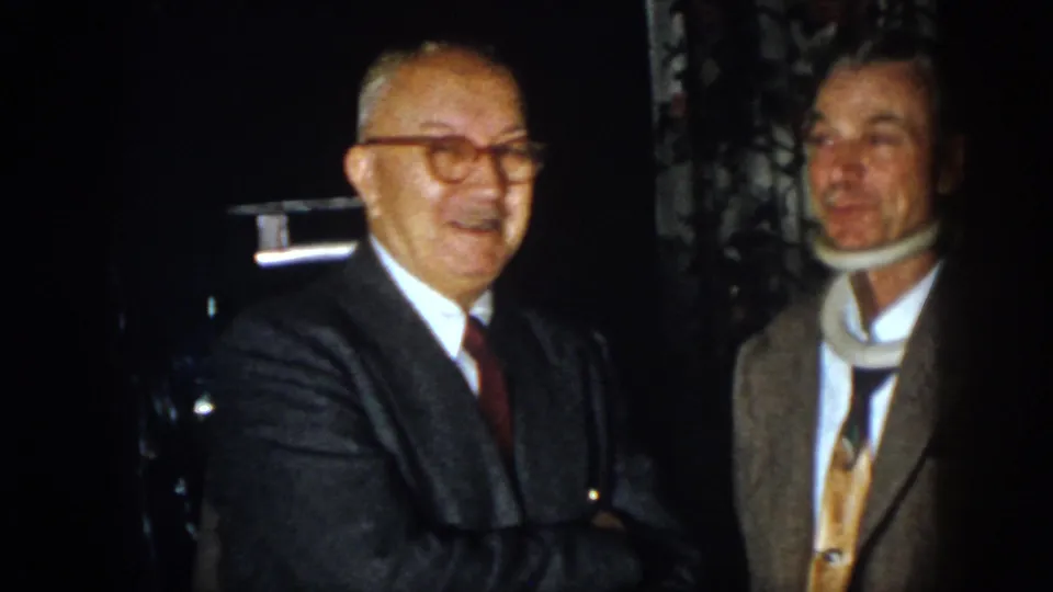 1958 Virginia frame with a man in glasses and a suited man wearing a neck brace in a dim indoor setting.