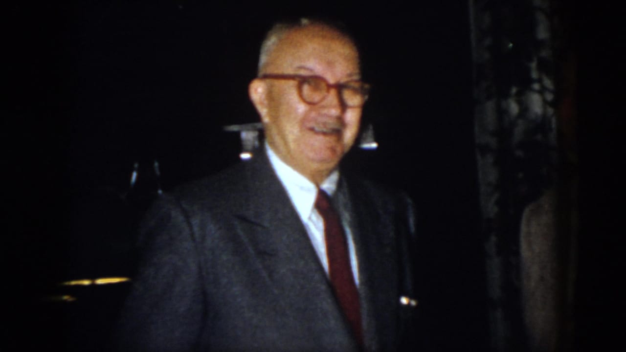 In 1958 Virginia home-movie footage, an older man in glasses, suit, and red tie smiles during a nighttime gathering.